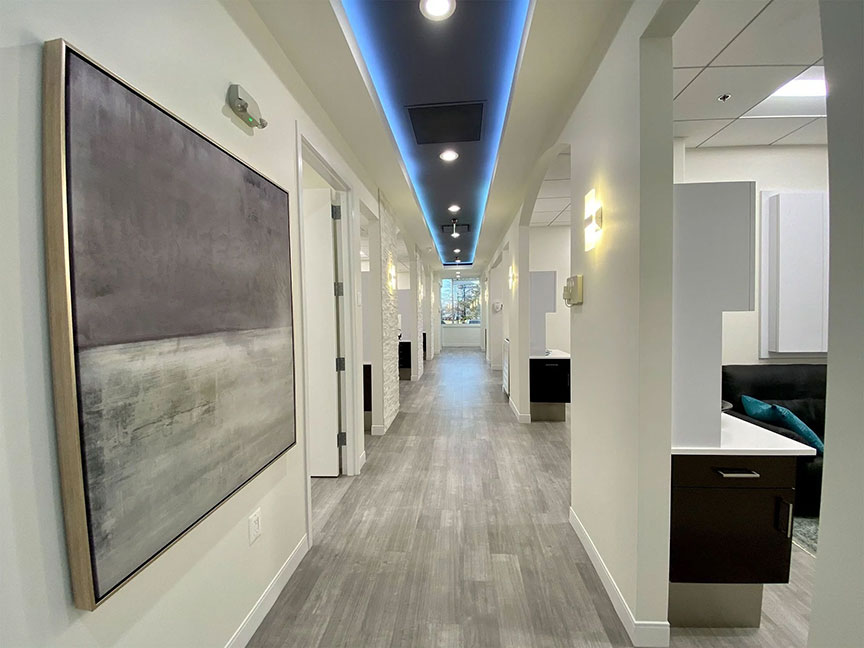 The image shows an interior hallway of a building, featuring a long corridor with a modern design, including a light wood floor, white walls, and contemporary fixtures.