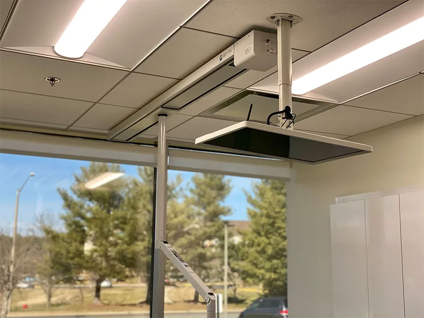 The image shows a ceiling-mounted projector with a screen extension, situated above a classroom or lecture hall setting.
