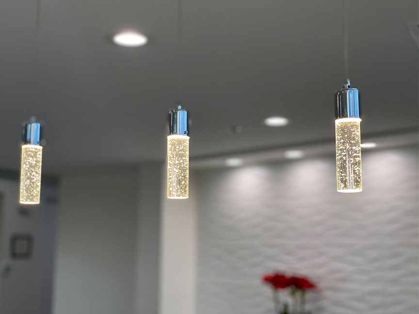 The image shows a modern interior space with an arrangement of three pendant lights hanging from the ceiling. Each light features a cylindrical design and is illuminated, casting a warm glow on the surrounding area. The background includes a white wall and what appears to be a decorative element or artwork on the right side.