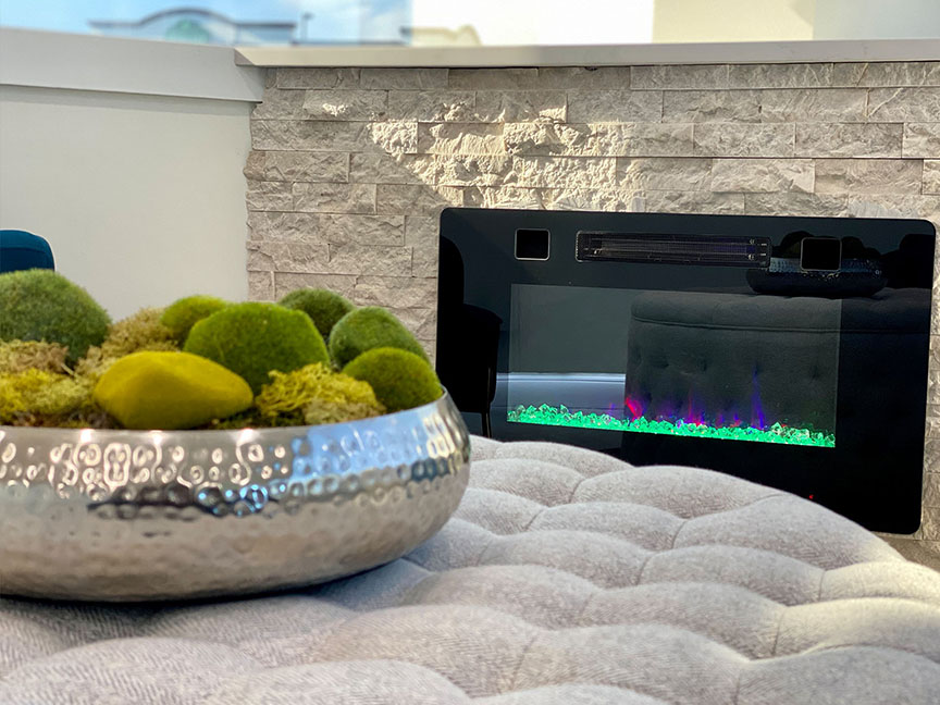 A modern electric fireplace with a sleek black frame, set into a beige brick wall, accompanied by a decorative vase with spherical green moss balls and a silver bowl filled with small round objects.