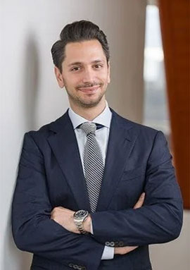 The image shows a man dressed in business attire, standing against a wall with his arms crossed.