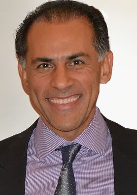 The image is a photograph of a smiling man with dark hair and facial stubble, wearing a suit. He appears to be at a formal event or in a professional setting.