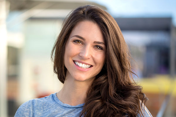 Smiling woman with long brown hair, wearing a gray top and standing outdoors in front of a building.