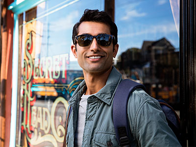 The image features a man standing in front of a store window, wearing sunglasses and a backpack, with a smile on his face.