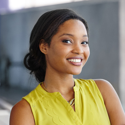 The image shows a woman with a bright smile, wearing a yellow top and a lanyard around her neck. She has her hair styled upwards and is standing against a backdrop of a building wall.