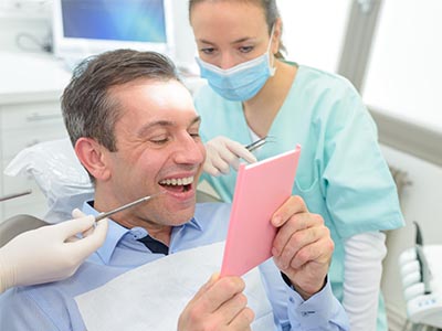 A dentist is holding a pink card while sitting in his chair, with a smiling woman standing behind him.