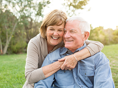 An elderly couple embracing outdoors, with a woman in a light-colored top and man in a dark shirt.