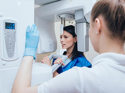 Woman in blue gown and white gloves standing next to a large, modern imaging machine.