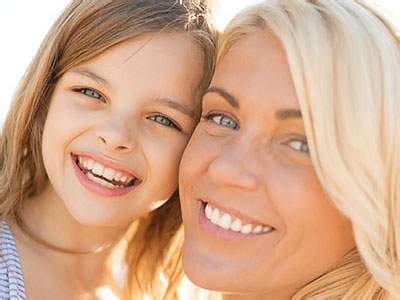 A woman and a young girl smiling at the camera, with the woman appearing to be in her late 30s or early 40s and the child around 6 years old.