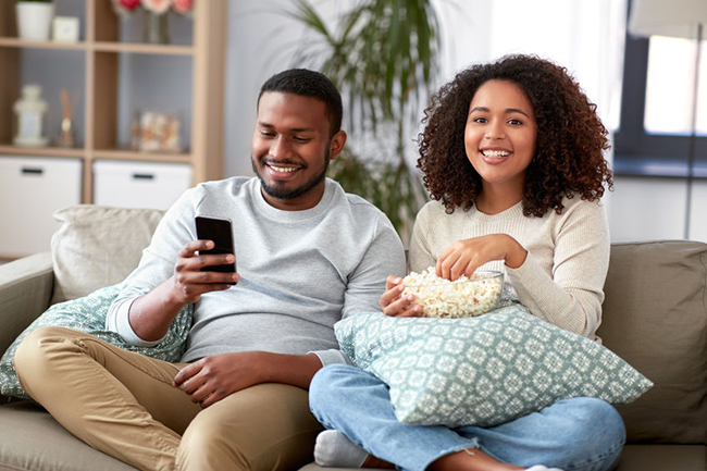 A man and woman sitting on a couch, sharing a moment of leisure while enjoying popcorn.