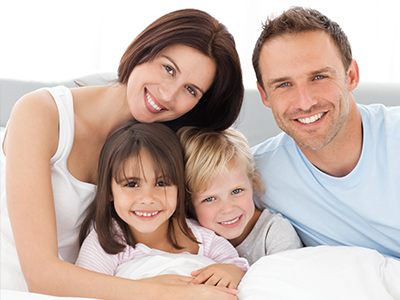 The image shows a family of four, including two adults and two children, smiling and posing together in bed.