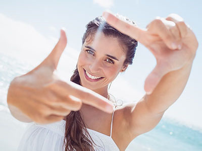 A woman takes a selfie with one hand, smiling and holding up her index finger against the bright sky.