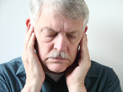 A man with a mustache, wearing glasses and a blue shirt, has his hand on his ear and appears to be in pain or discomfort.